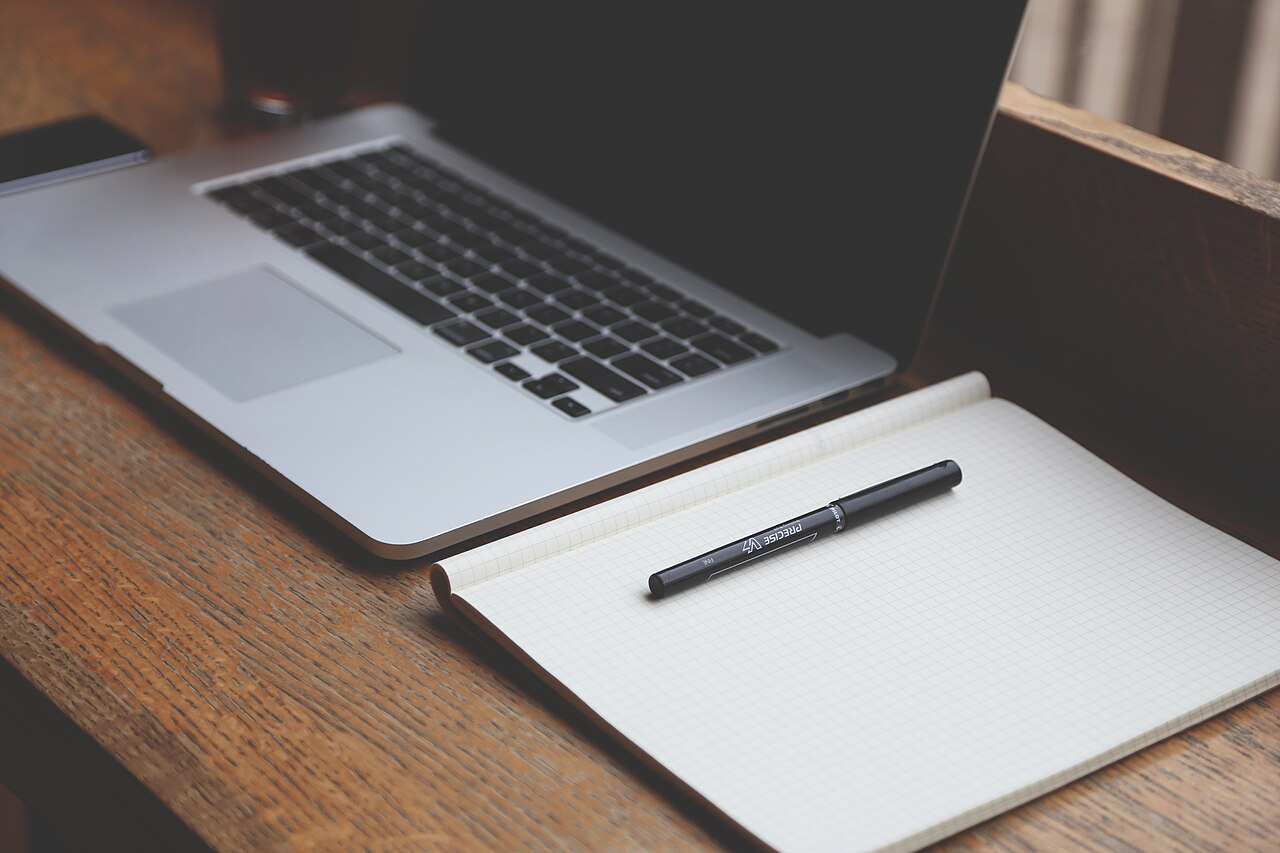 An open laptop with a blank screen beside a notebook and pen on a wooden table.