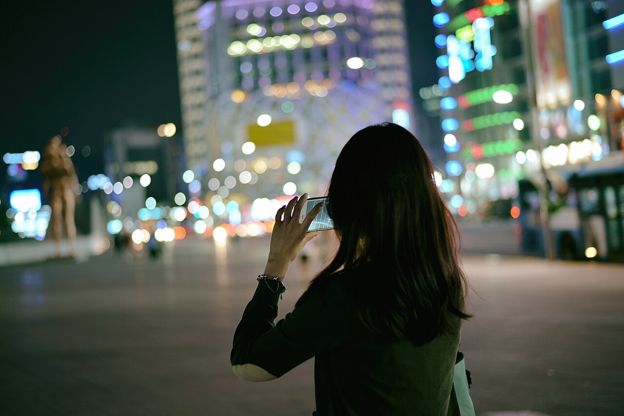 A person seen from behind uses a smartphone amid blurred city lights in Seoul at night.