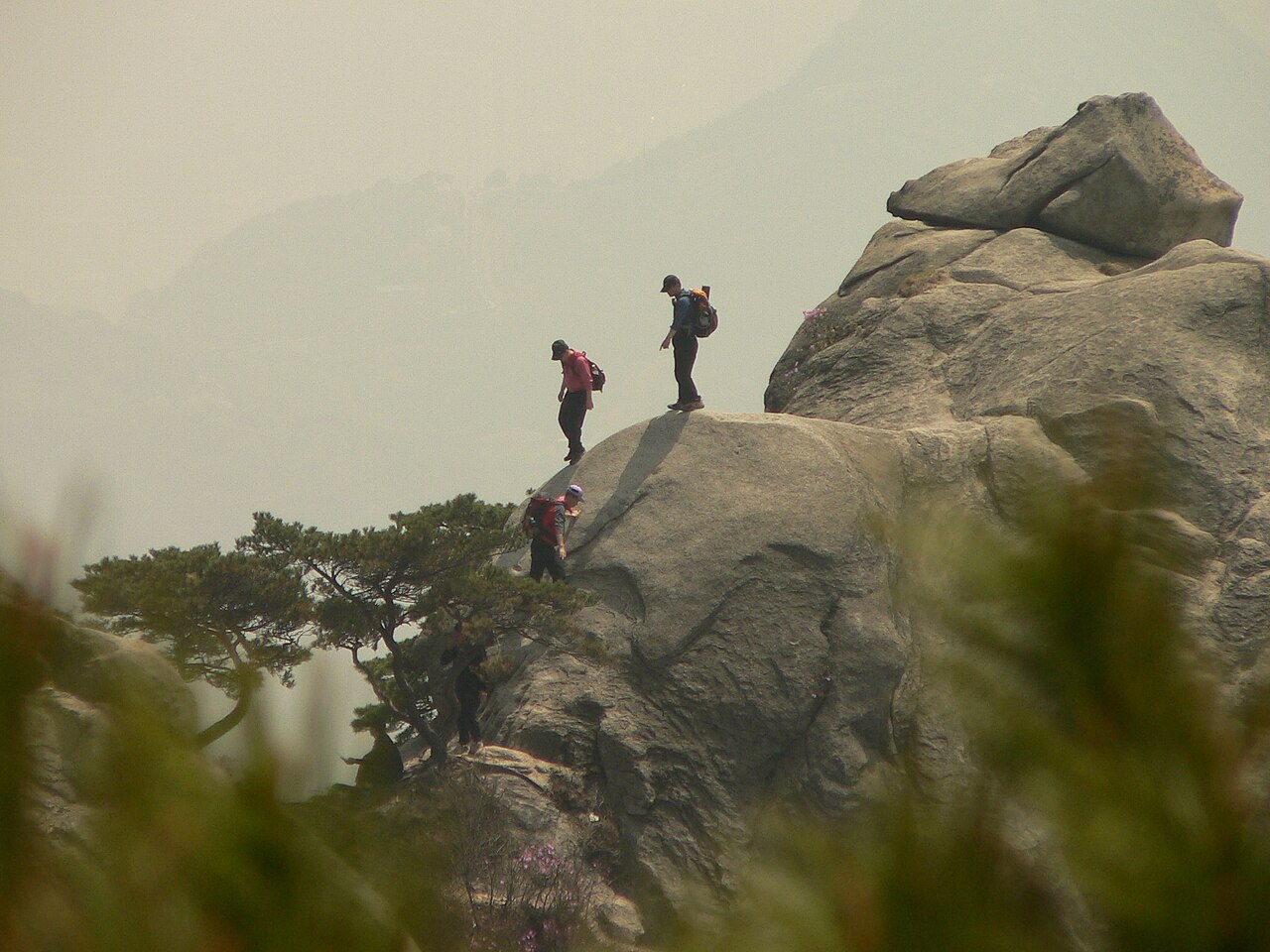 Three hikers in bright red jackets with backpacks climbing a large granite rock at Bukhansan in hazy weather.