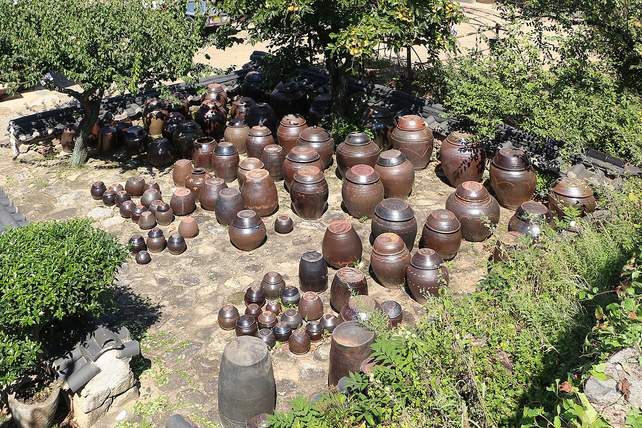Dozens of dark brown onggi jars of different sizes standing on a stone terrace surrounded by greenery.